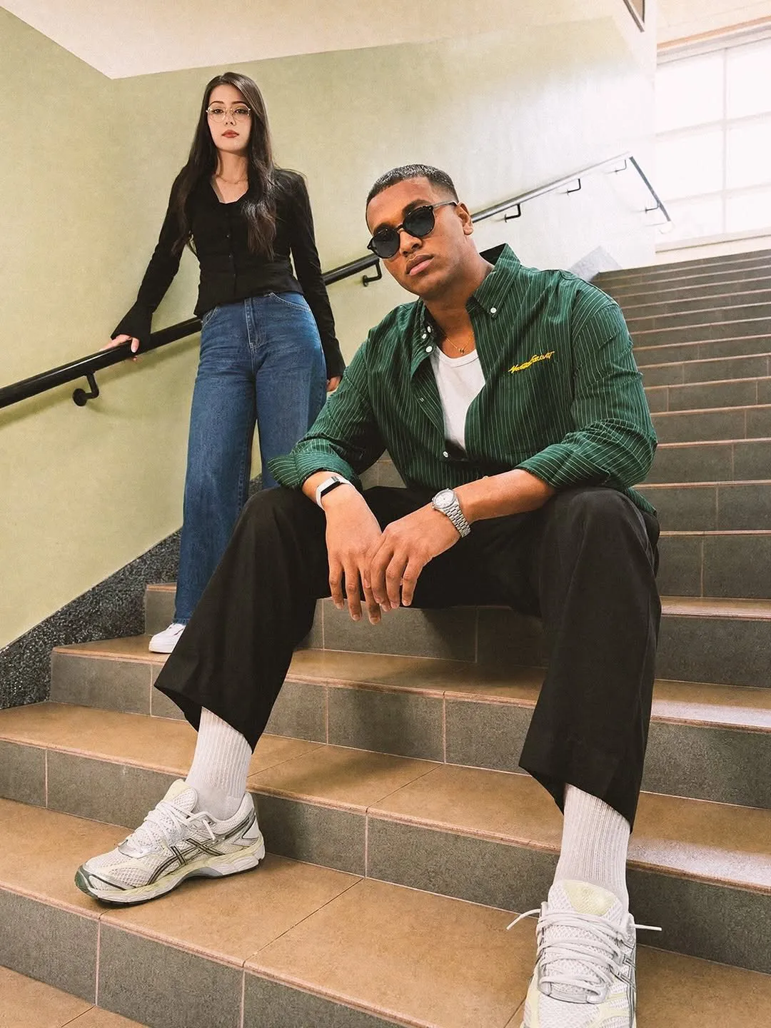 Man in round black sunglasses on a staircase, a woman in fine round optical frames standing behind him
